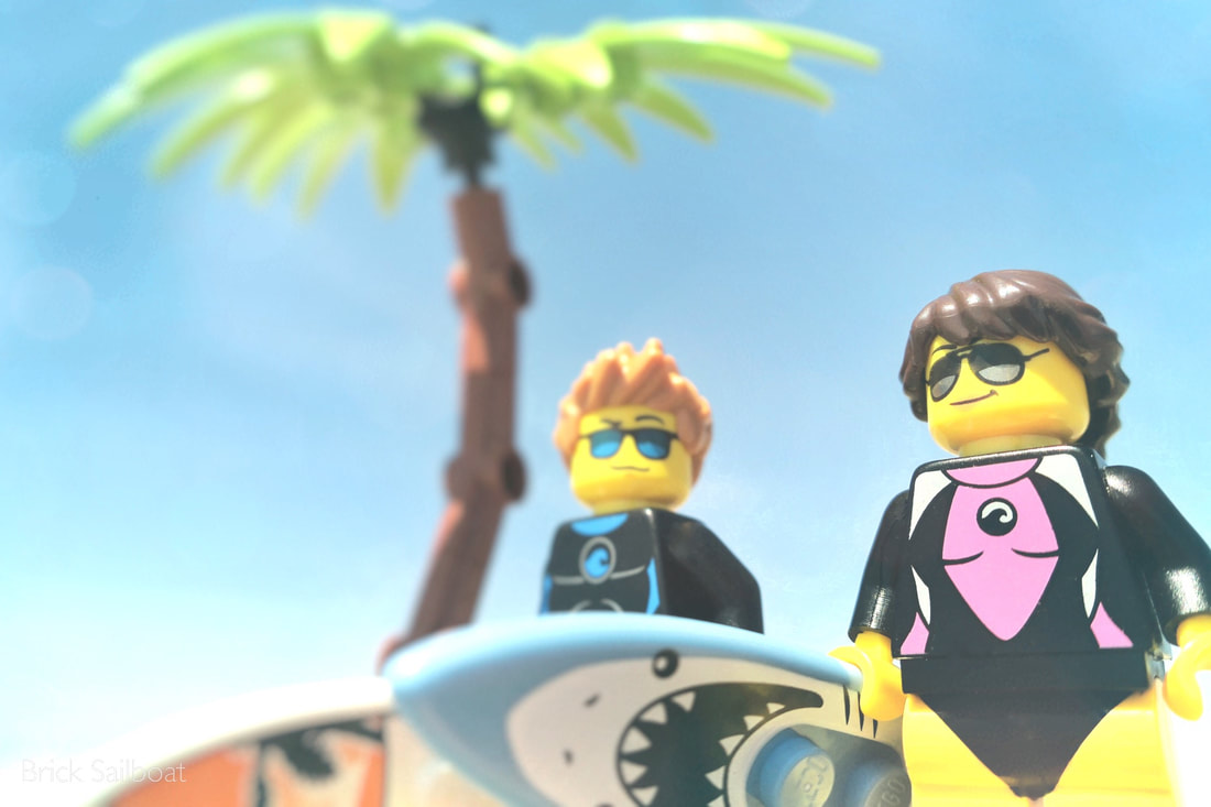 A couple of LEGO surfers on the beach under a palm tree.
